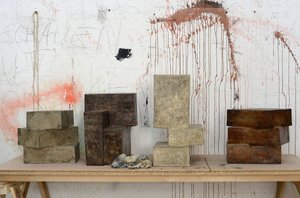 The color photograph shows four concrete sculptures in different shades of grey by Rouven Dürr, each composed of two to three cuboids. They stand on a simple table consisting of a wooden top and trestles. The studio wall in the background shows splashes of paint, the word “schauen” [look] written in delicate capitals is recognizable.