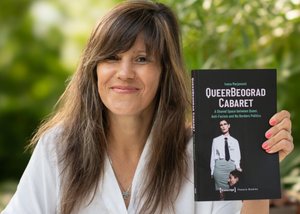 The color photograph in landscape format shows the smiling author Ivana Marjanović. She wears her medium-length brown hair loose and parted on the left side, with long fringes covering her forehead. In her left hand she is holding the recently published book about the QueerBeograd Cabaret. Her nails are painted salmon color. Blurred foliage can be seen in the background.